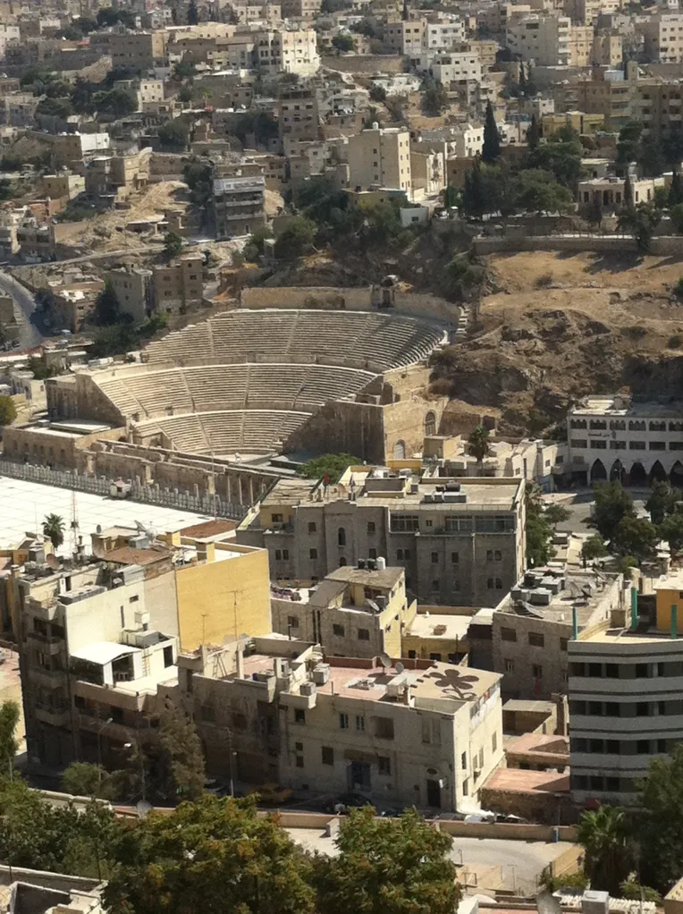 Aerial view of a hillside city with dense sand-colored buildings surrounding an ancient stone amphitheater, warm earthy tones and a reflective, timeless mood
