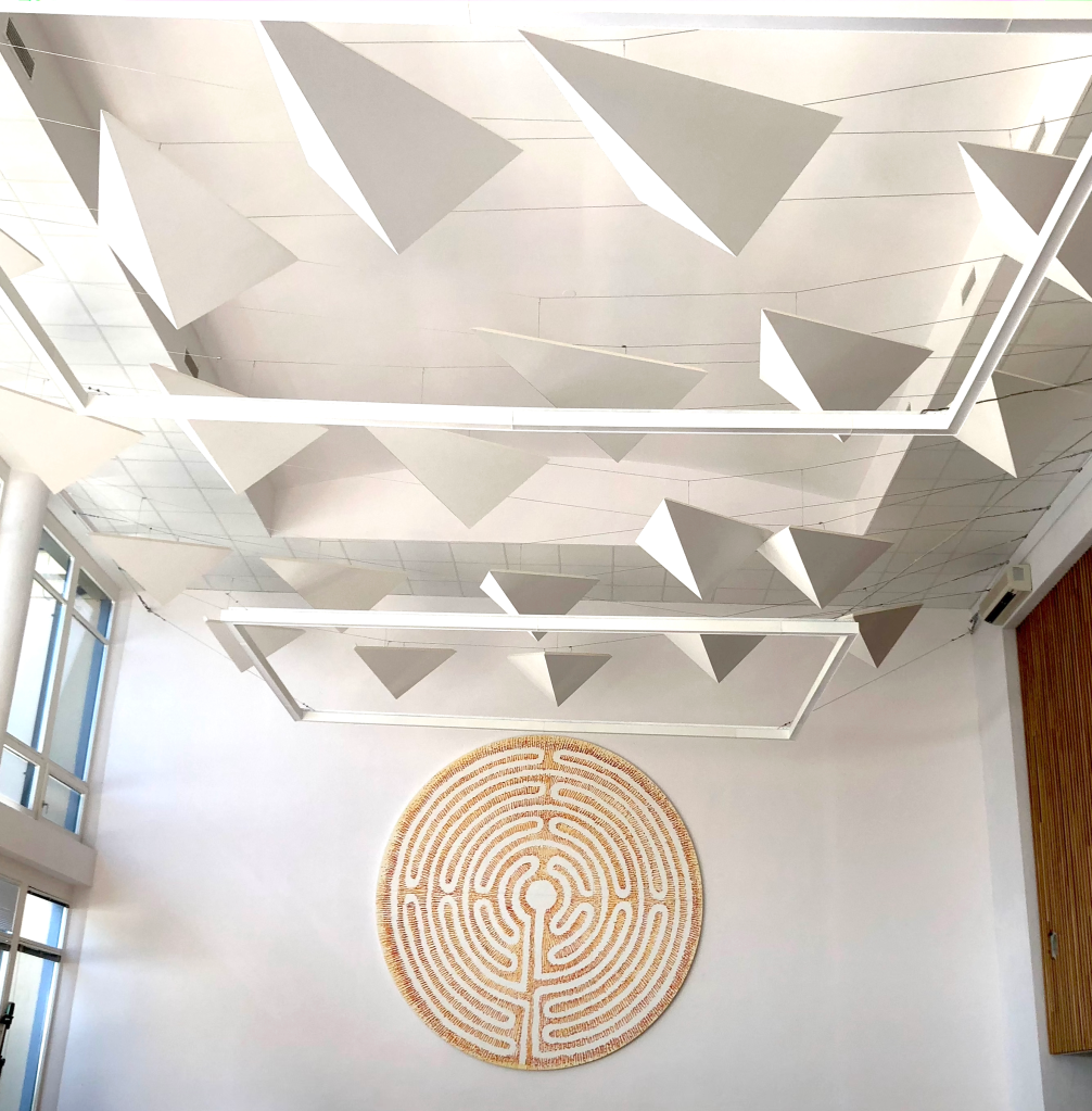 Interior ceiling with hanging geometric triangular panels suspended on wires above a circular labyrinth artwork, lit in warm teal and navy tones with a calm, thoughtful mood
